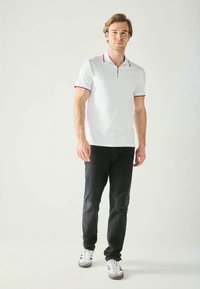 Vit polo med dragkedja och röda detaljer vid kragen, kombinerad med mörka jeans och vita sneakers, bärs av en stående manlig figur.