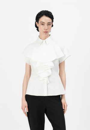 White shirt with a large, cascading ruffle down the front, short sleeves, and a button-down collar, paired with black pants.