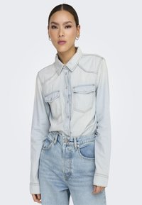 Camisa de mezclilla azul claro con mangas largas, cuello puntiagudo, botones a presión, dos bolsillos frontales y costuras en contraste, combinada con jeans azul claro.
