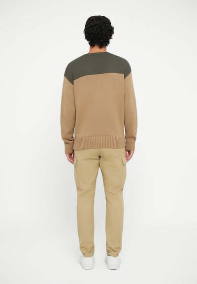 Suéter de punto en tonos marrón claro y verde oscuro, con un diseño a bloques de color y un dobladillo acanalado. Combinado con pantalones cargo beige y zapatillas blancas.