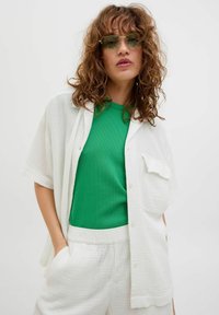 Camisa blanca de textura grande y oversize con mangas cortas sobre una camiseta verde de canalé. Pantalones cortos blancos con cintura elástica. La modelo lleva gafas de sol con tintado verde.
