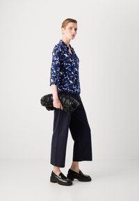 Μπλούζα ναυτικού μπλε με μοτίβο και τρία τέταρτα μανίκια, συνδυασμένη με καφέ παντελόνι φαρδύ και χοντρές μαύρες loafers, κρατώντας μια υφασμάτινη μαύρη τσάντα.