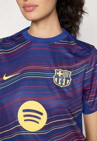 Blå sportströja med flerfärgade horisontella ränder, prydd med en gul Spotify-logga och FC Barcelonas märke på framsidan, tillverkad av mjukt tyg.