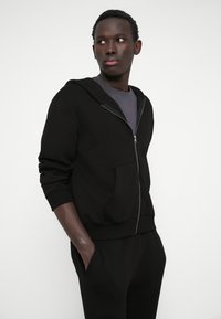 Black zip-up hoodie in cotton blend with a textured finish, featuring a front pocket and ribbed cuffs, worn over a gray T-shirt.