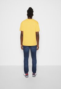 Yellow cotton t-shirt with short sleeves, paired with blue denim jeans and white sneakers featuring red and blue accents, viewed from behind.