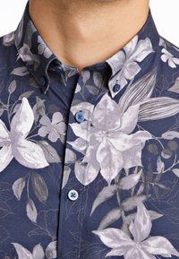 Camisa azul marino con estampado floral grande de flores claras, hojas verdes y botones azules; tela texturizada con detalle de cuello abotonado.