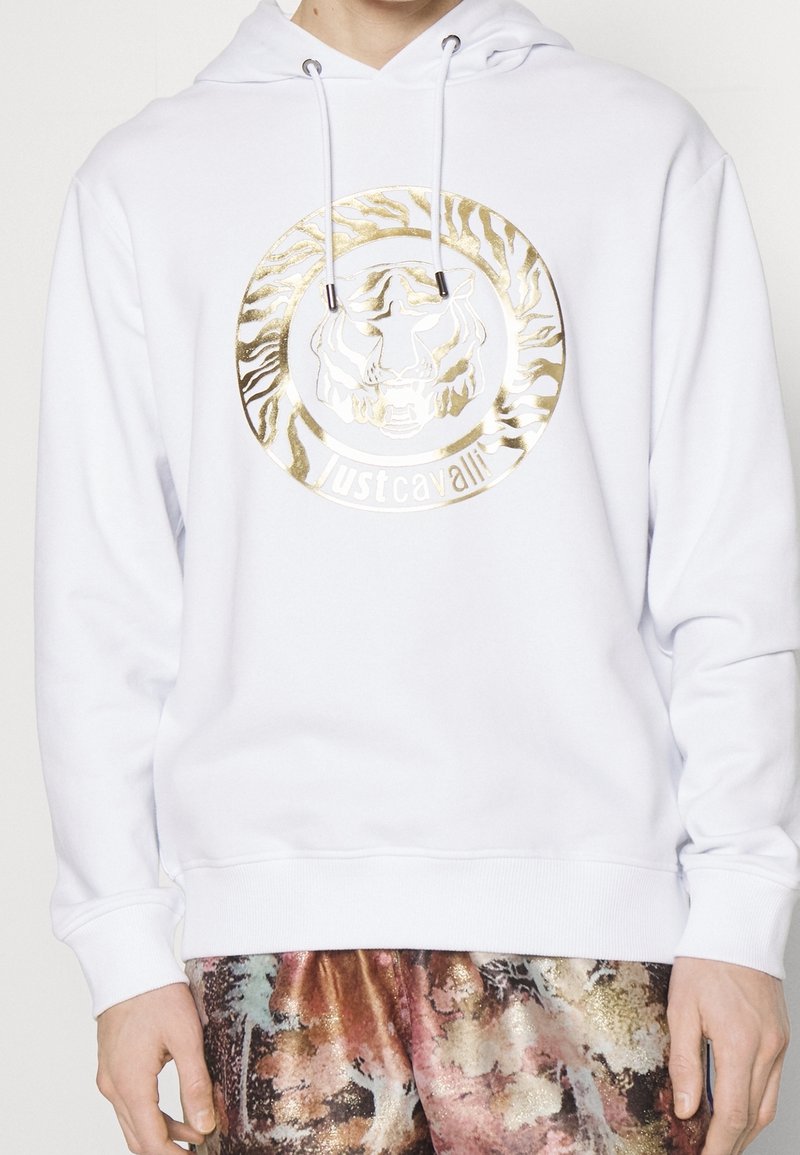 Sweat à capuche blanc avec un visage de tigre doré brillant et le texte « Just Cavalli » sur la poitrine, porté avec un pantalon multicolore à motifs.