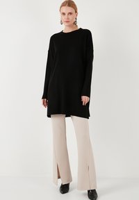 Tunique nera a coste con maniche lunghe, abbinata a pantaloni beige a gamba larga con spacchi laterali. Stivaletti neri completano l'outfit.