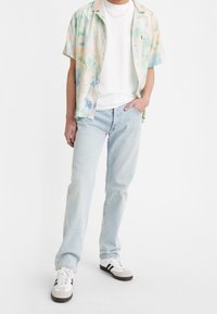 Kortärmad, knappad skjorta i pastellfärger med abstrakta mönster, ihop med ljusblå jeans och vita sneakers med svarta accenter.