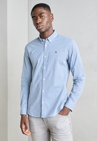 Blue and white checkered long-sleeve shirt with a button-down collar and small logo on the chest, paired with light gray jeans.