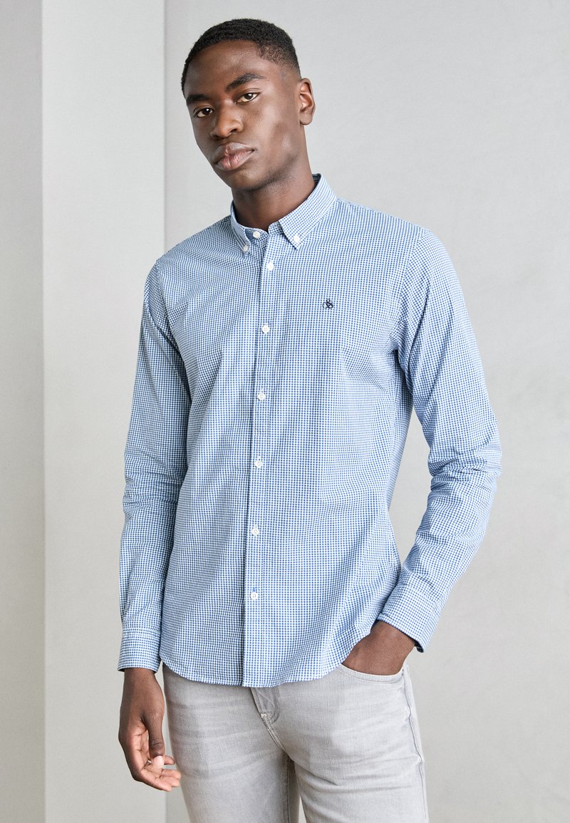 Blue and white checkered long-sleeve shirt with a button-down collar and small logo on the chest, paired with light gray jeans.