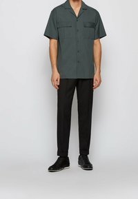 Kratší rukáv tmavě zelené košile se dvěma kapsami na hrudi, černé knoflíky; ve spojení s černými kalhotami a černými teniskami. Hladká textura látky.