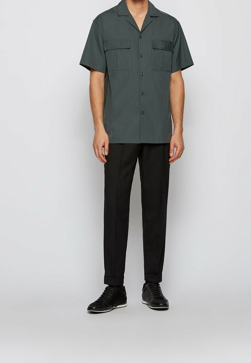 Kratší rukáv tmavě zelené košile se dvěma kapsami na hrudi, černé knoflíky; ve spojení s černými kalhotami a černými teniskami. Hladká textura látky.