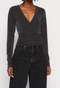 Svart långärmad topp med djup V-ringning, har en skimrande textur och åtsittande design. Kombinerad med högmidjade svarta jeans.