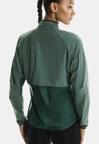 Jachetă sport verde, realizată dintr-un material ușor și rezistent la apă, cu mâneci raglan și texturi contrastante pe secțiunile superioare și inferioare.