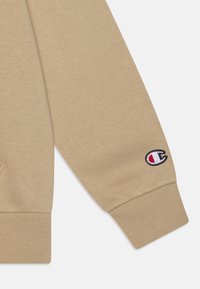 Vaaleanbeige-hupparihaalari, joka on valmistettu pehmeästä kankaasta. Hihassa on joustava resori ja siinä on pieni brodeerattu logo, jossa on punaisia, valkoisia ja sinisiä yksityiskohtia.