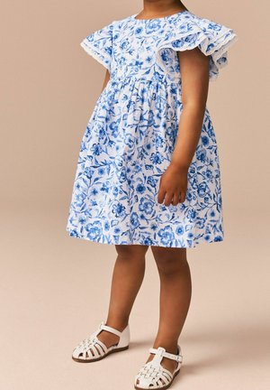 Vestito floreale blu e bianco con maniche arricciate, corpino aderente e gonna svasata. Abbinato a sandali bianchi con cinturini.