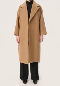 Camel-wollmantel mit lockerer Passform, breiten Revers, doppelter Knopfleiste und seitlichen Bindebändern. Über einem schwarzen Rollkragenpullover und schwarzen Hosen getragen.