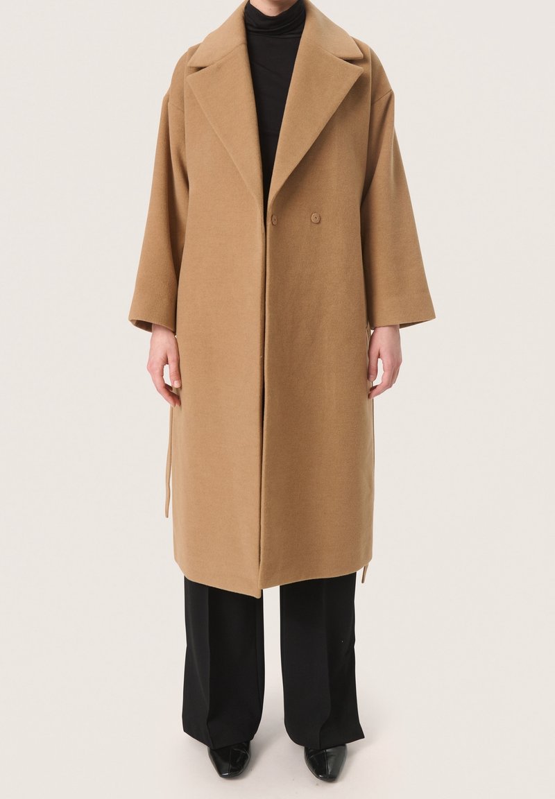 Camel-wollmantel mit lockerer Passform, breiten Revers, doppelter Knopfleiste und seitlichen Bindebändern. Über einem schwarzen Rollkragenpullover und schwarzen Hosen getragen.