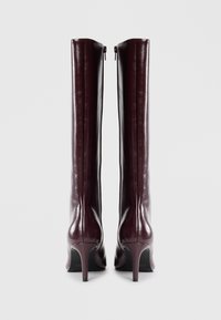 Une paire de bottes en cuir bordeaux foncé, hautes, avec des talons fins et des fermetures éclair à l'arrière, se tenant côte à côte sur un fond clair.