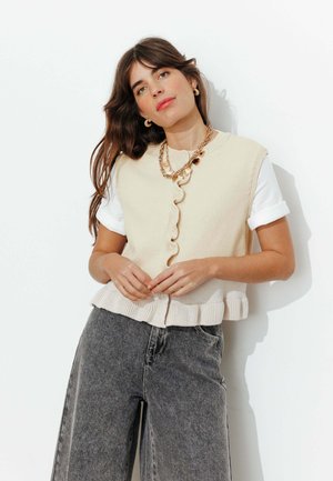 Femme portant un gilet sans manches en maille crème par-dessus une chemise blanche à manches retroussées et un jean gris taille haute à jambes larges, posant devant un mur blanc.