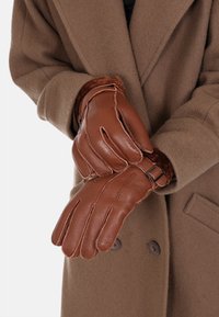 Guanti in pelle marrone con un morbido rivestimento in pelliccia, caratterizzati da un accentuato con fibbia e dettagli cuciti, indossati sotto un cappotto marrone.