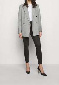 Blazer gris cruzado con doble botonadura, tejido texturizado, botones negros, combinado con leggings negros y tacones negros de punta afilada. Camiseta blanca sencilla debajo.