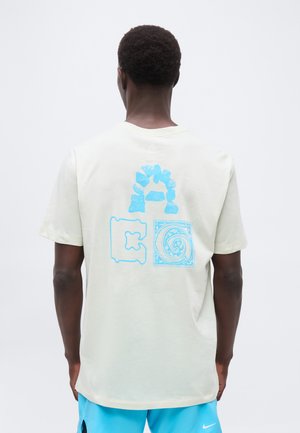 Homme portant un t-shirt blanc avec trois impressions artistiques de lettres bleues dans le dos, associé à un short Nike bleu vif.