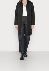 Svart vadderad lång kappa med dragkedja, bärs över en vit tröja och mönstrad skjorta, ihop med hög midja mörka jeans och svarta stövlar.