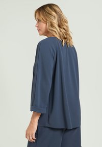 Mujer rubia con cabello ondulado, vistiendo una blusa azul marino de manga tres cuartos suelta y pantalones a juego, mirando hacia la izquierda.