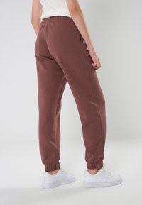 Braune Sweatpants mit elastischem Bund und Bündchen, ausgestattet mit Seitentaschen. Kombiniert mit weißen Turnschuhen vor einem neutralen Hintergrund.