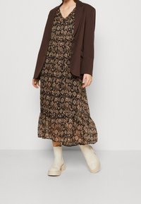Blazer marron porté sur une robe midi à motifs dans des tons foncés, avec un ourlet à volants et des bottes à semelle épaisse. Tissu texturé et silhouette définie.