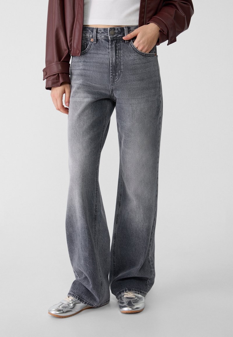 Jean large en denim gris avec une finition délavée, taille haute et design à cinq poches, associé à des chaussures argentées et une veste marron.