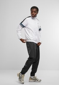 Vit zip-up jacka med svarta och blå axelränder, kombinerad med grå byxor. Sneakers har en design i silver och guld.