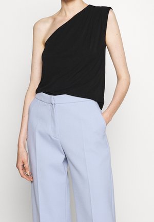 Kobieta w czarnej, bezrękawnej, asymetrycznej bluzce wsuniętej w wysokie, jasnoniebieskie, dopasowane spodnie, stojąca z jedną ręką w kieszeni.