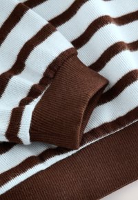 Detalle de suéter de punto con rayas marrones y blancas, que presenta una textura suave y puños marrones acanalados. Las rayas están distribuidas uniformemente.