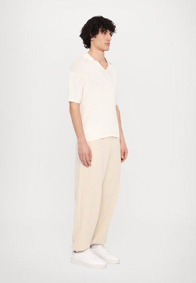 Pletené polo tričko s krátkym rukávom v off-white farbe s výstrihom do V, v kombinácii so svetlobežovými nohavicami a bielymi teniskami, zobrazené z boku.