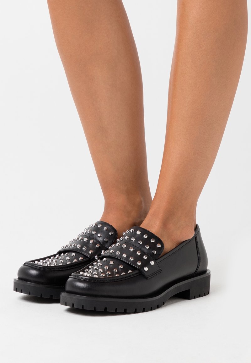 Des mocassins noirs présentent un design clouté sur le upper avec deux lanières, une semelle épaisse et une texture en cuir lisse.