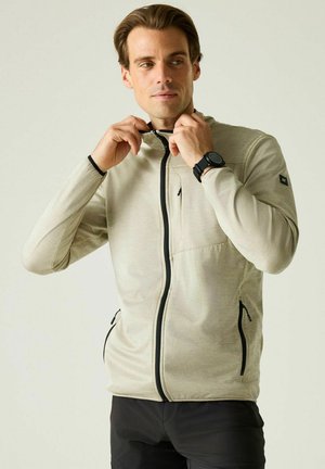 Hombre ajustándose el cuello de una chaqueta beige con cremallera negra, lleva un reloj de pulsera negro y pantalones negros, mirando ligeramente hacia un lado.