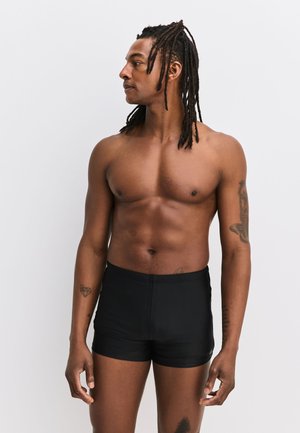 Muskelbepackter Mann mit langen Dreadlocks, der schwarze Badehose trägt, seitlich stehend vor einem einfarbigen hellgrauen Hintergrund.
