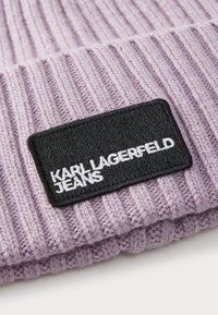 Lilac ribbed knit beanie featuring a black rectangular patch with white text "KARL LAGERFELD JEANS." Textured fabric with a fold at the bottom.