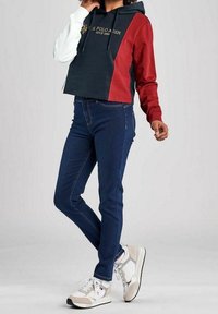 Person som bär en hoodie i tre färger med ärmar i marinblått, vitt och rött, mörkblå jeans och vita sneakers, stående med ett ben korsat.