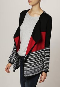 Cardigan nero e rosso con un motivo a righe. Presenta una scollatura aperta, maniche lunghe e un tessuto a maglia testurizzato, abbinato a una maglietta grigia sotto.