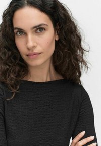 Czarna, teksturowana bluzka z długim rękawem i dekoltem w łódkę. Modelka ma kręcone włosy i neutralny wyraz twarzy, z widocznymi detalami skóry.