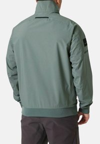 Chaqueta verde ligera con un alto cuello, puños acanalados y dobladillo. Presenta una textura sutil y un acento rectangular en la manga.