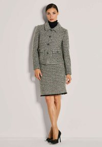 Frau, die an einer Wand steht und einen grauen Tweed-Blazer sowie einen passenden Rock trägt, dazu schwarze High Heels und einen schwarzen Rollkragenpullover.