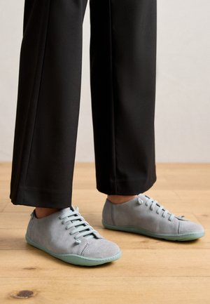 Personne portant des baskets en cuir suédé gris clair avec des semelles vertes et un pantalon noir, se tenant sur un sol en bois.