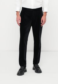 Zwarte fluwelen broek met een getailleerde pasvorm, met een soepele textuur en voorplooien. Gecombineerd met zwarte instappers en een wit shirt.
