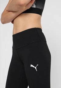 Černé legíny vyrobené z elastického materiálu, s vysokým pasem a bílým logem Puma na levém stehně. Hladký povrch a těsný střih.