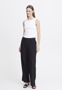Weißes ärmelloses Oberteil, schwarze locker sitzende Hose, schwarze Slippers. Glatter Stoff, schlichtes Design und klare Linien.